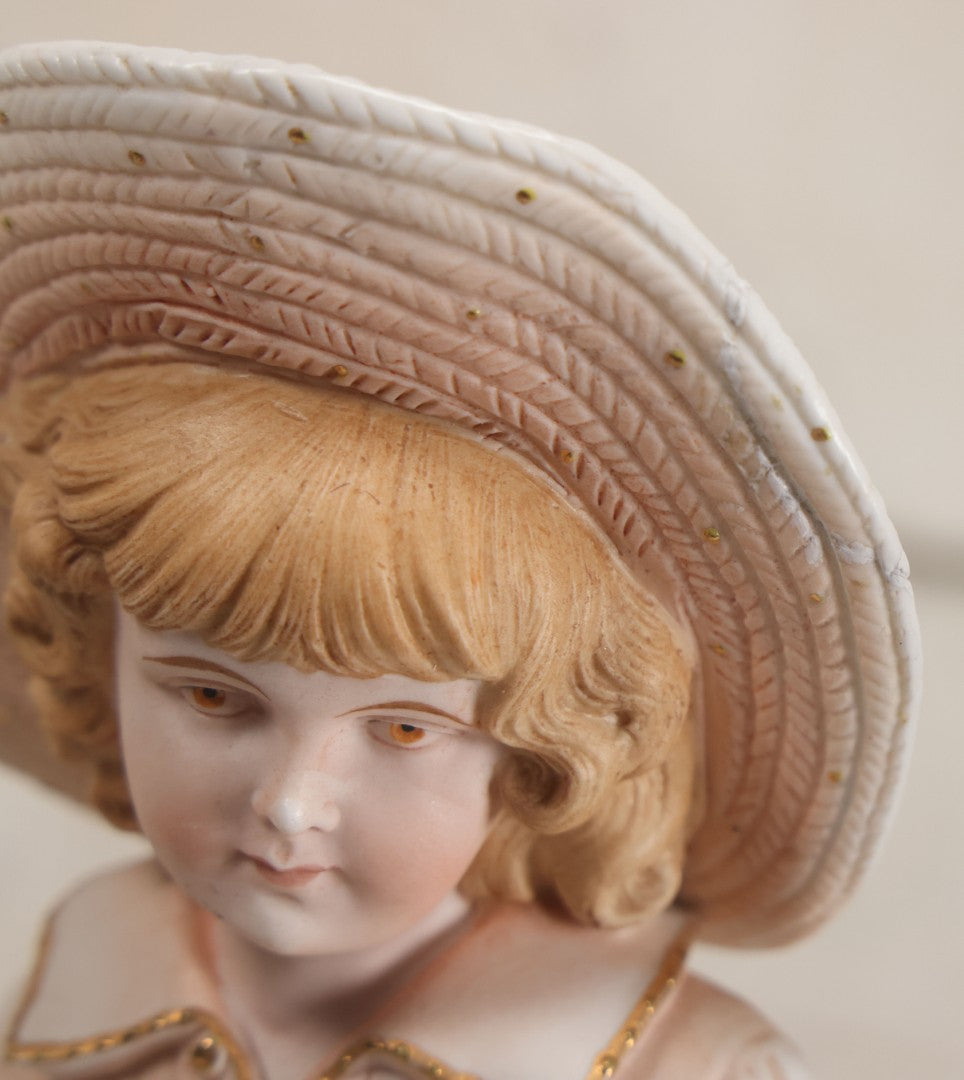 Lot 037 - Vintage Bisque Statue Of Young Blond Child In Large Straw Hat Sitting By Shore With Pail, With Pink Painted Details And Gold Embellishments, Note Chip, Repairs, 12" Height