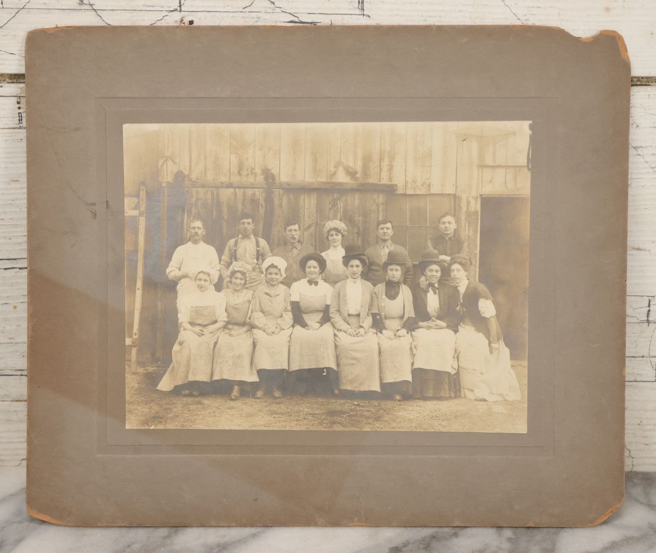 Lot 142 - Antique Boarded Occupational Photograph Of Men And Women Workers Posing In Workwear Outside A Barn, Women In Bowler Hats And Bonnets And Man Smoking Pipe