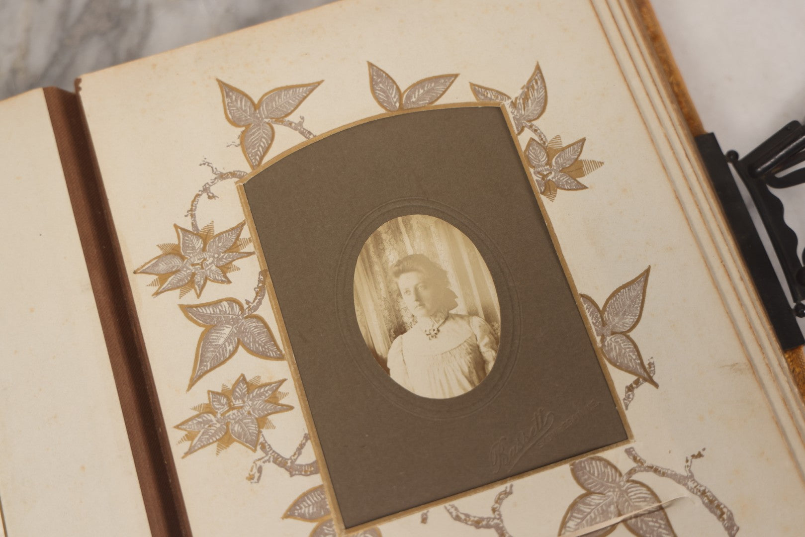 Lot 012 - Antique Victorian Photo Album With Wood Cover And Silverplate Emblem, Containing Twenty Antique Photographs Of Various Types, Including Man In Office, Children With Pram, Man With White Beard, Note Binding Wear Cover Loosening