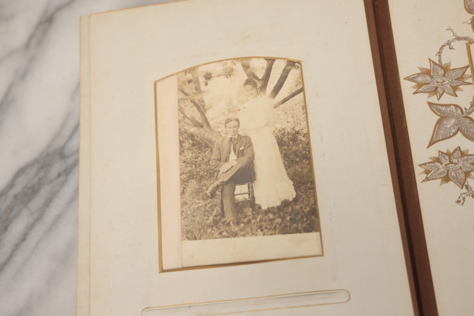 Lot 012 - Antique Victorian Photo Album With Wood Cover And Silverplate Emblem, Containing Twenty Antique Photographs Of Various Types, Including Man In Office, Children With Pram, Man With White Beard, Note Binding Wear Cover Loosening