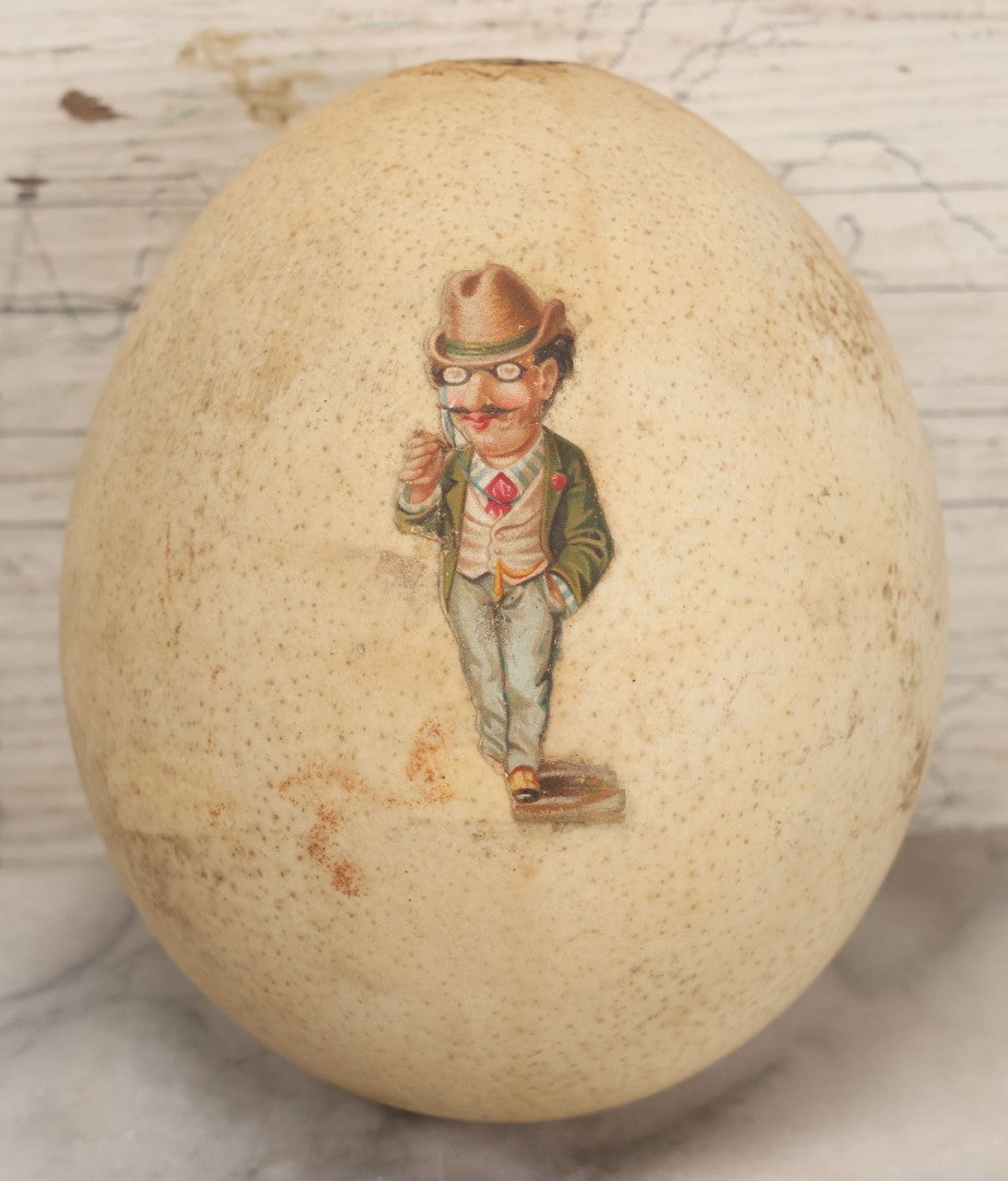 Lot 017 - Antique Genuine Ostrich Egg Specimen With Two Applied Victorian Die Cut Scraps Of Cheeky Caricatures, Note Originally Had More Scraps Attached, As Found