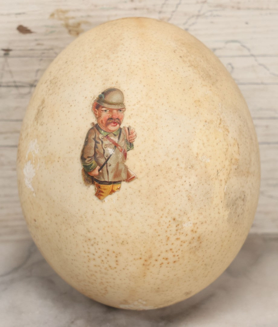 Lot 017 - Antique Genuine Ostrich Egg Specimen With Two Applied Victorian Die Cut Scraps Of Cheeky Caricatures, Note Originally Had More Scraps Attached, As Found