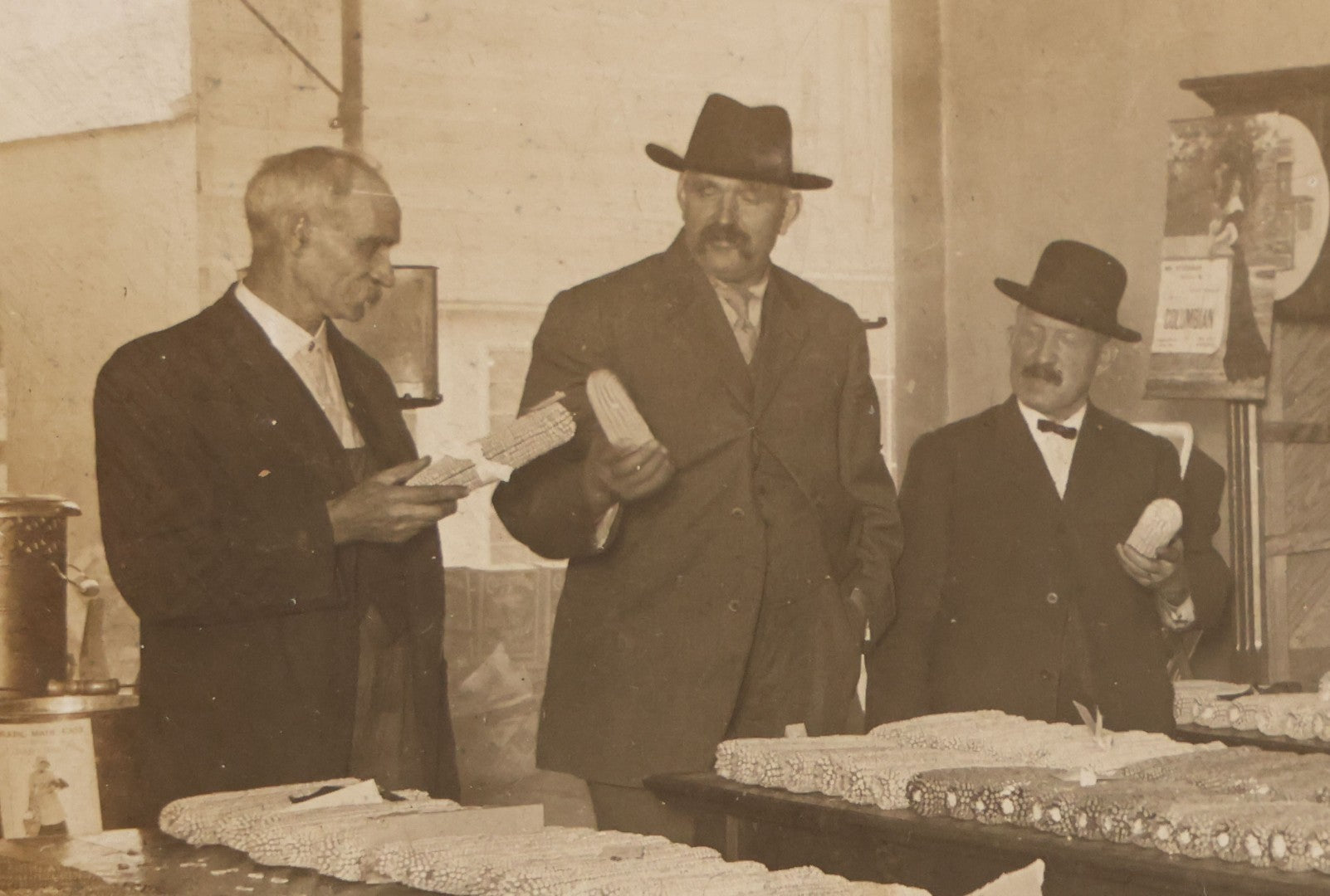Lot 084 - Antique Boarded Occupational Photograph Of Three Men Inspecting A Large Variety Of Corn, With Advertisement For "The 'Faultless' Malleable Iron Range" In Background