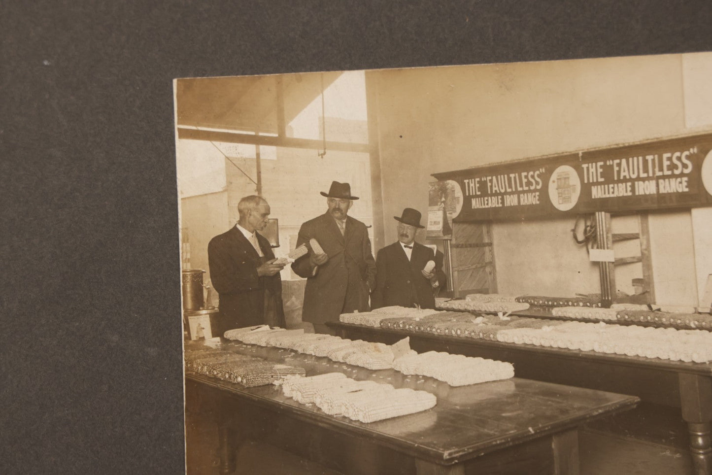 Lot 084 - Antique Boarded Occupational Photograph Of Three Men Inspecting A Large Variety Of Corn, With Advertisement For "The 'Faultless' Malleable Iron Range" In Background