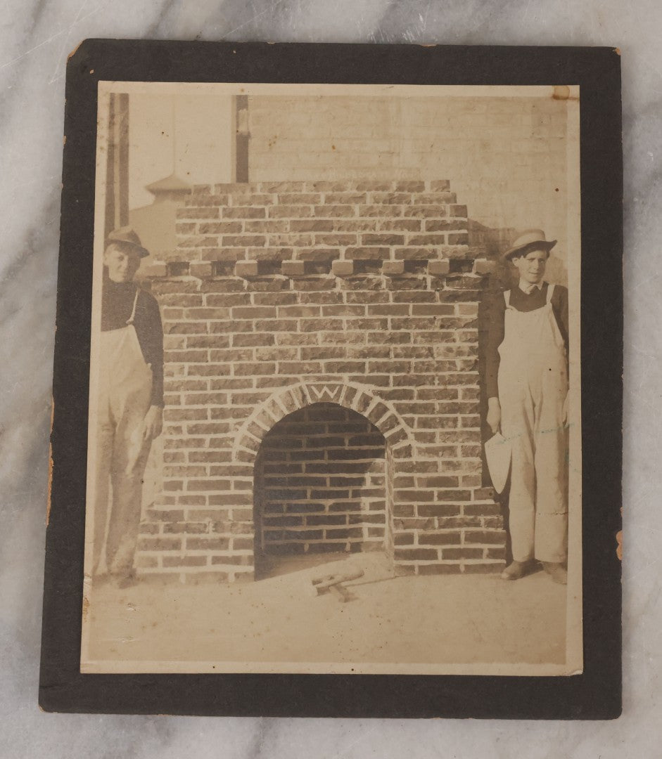 Lot 091 - Antique Boarded Photograph Of Two Young Bricklayers Posing With Tools In Front Of Newly Finished Fireplace With "W" Monogram