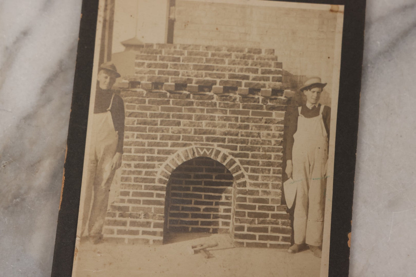 Lot 091 - Antique Boarded Photograph Of Two Young Bricklayers Posing With Tools In Front Of Newly Finished Fireplace With "W" Monogram