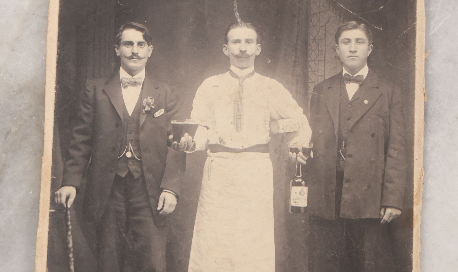 Lot 100 - Antique Trimmed Boarded Photograph Of Bartender Posing With Liquor Bottle, Glasses, And Cigar Box, With Two Men, Detailed Writing On Verso Likely In Polish