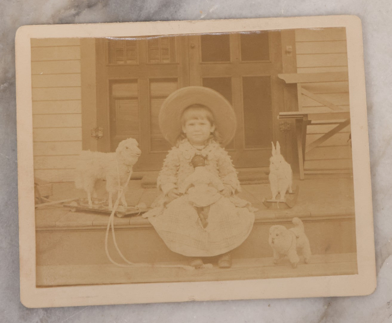 Lot 094 - Antique Boarded Photograph Of A Little Girl Posing On Porch With Her Toys: A Black Doll, Lamb Pull Toy, Dog Pull Toy, And Bunny Rocker, 
