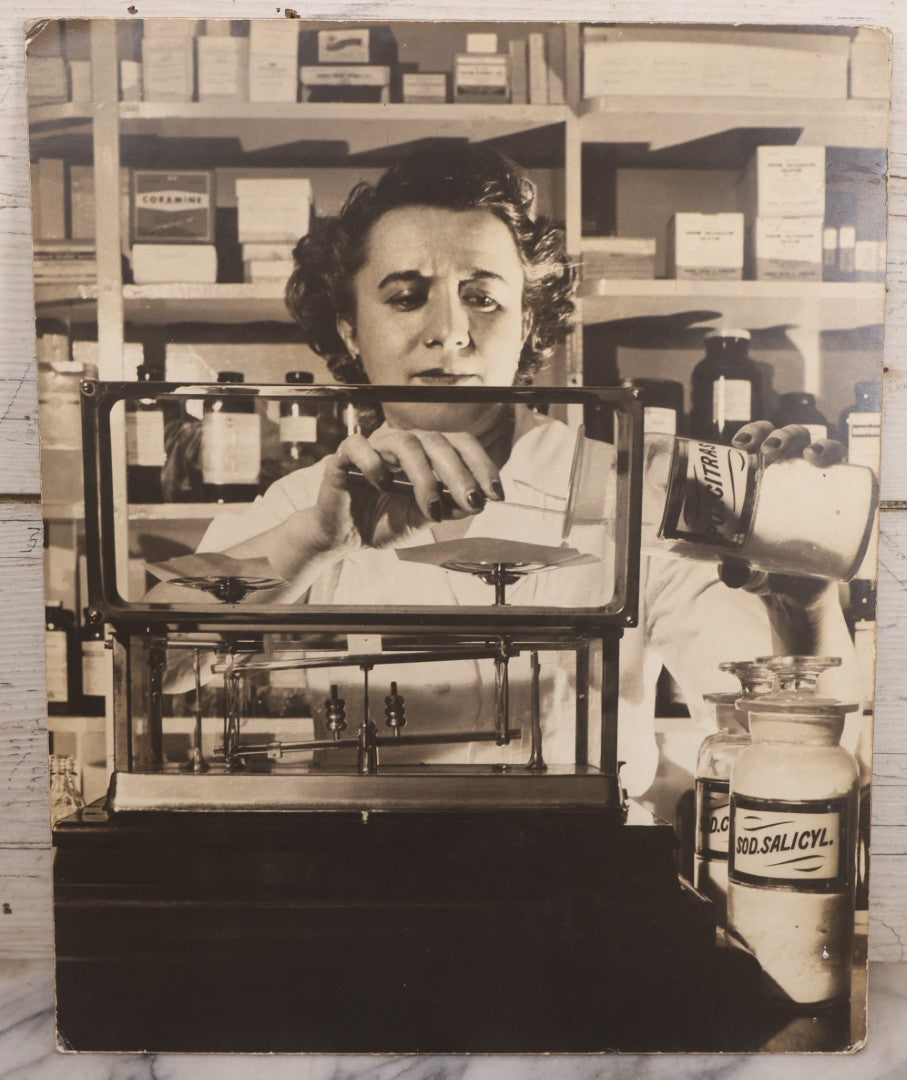 Lot 144 - Vintage Large Cardboard Mount Black And White Photograph Print Of Woman Pharmacist Measuring Drugs From Antique Glass Label Apothecary Jar, Many Drugs On Shelves In Background, 13-1/4" x 16-1/2"