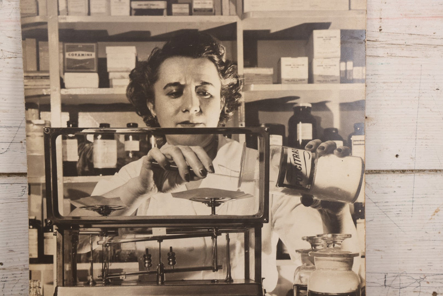 Lot 144 - Vintage Large Cardboard Mount Black And White Photograph Print Of Woman Pharmacist Measuring Drugs From Antique Glass Label Apothecary Jar, Many Drugs On Shelves In Background, 13-1/4" x 16-1/2"