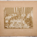 Lot 069 - Antique Boarded Photograph Of An Integrated Elementary School Class, Posing With Large 48 Star American Flag Bunting And Patriotic Decor, Pennants From P.S. 73, Teacher In Photo, Circa 1895