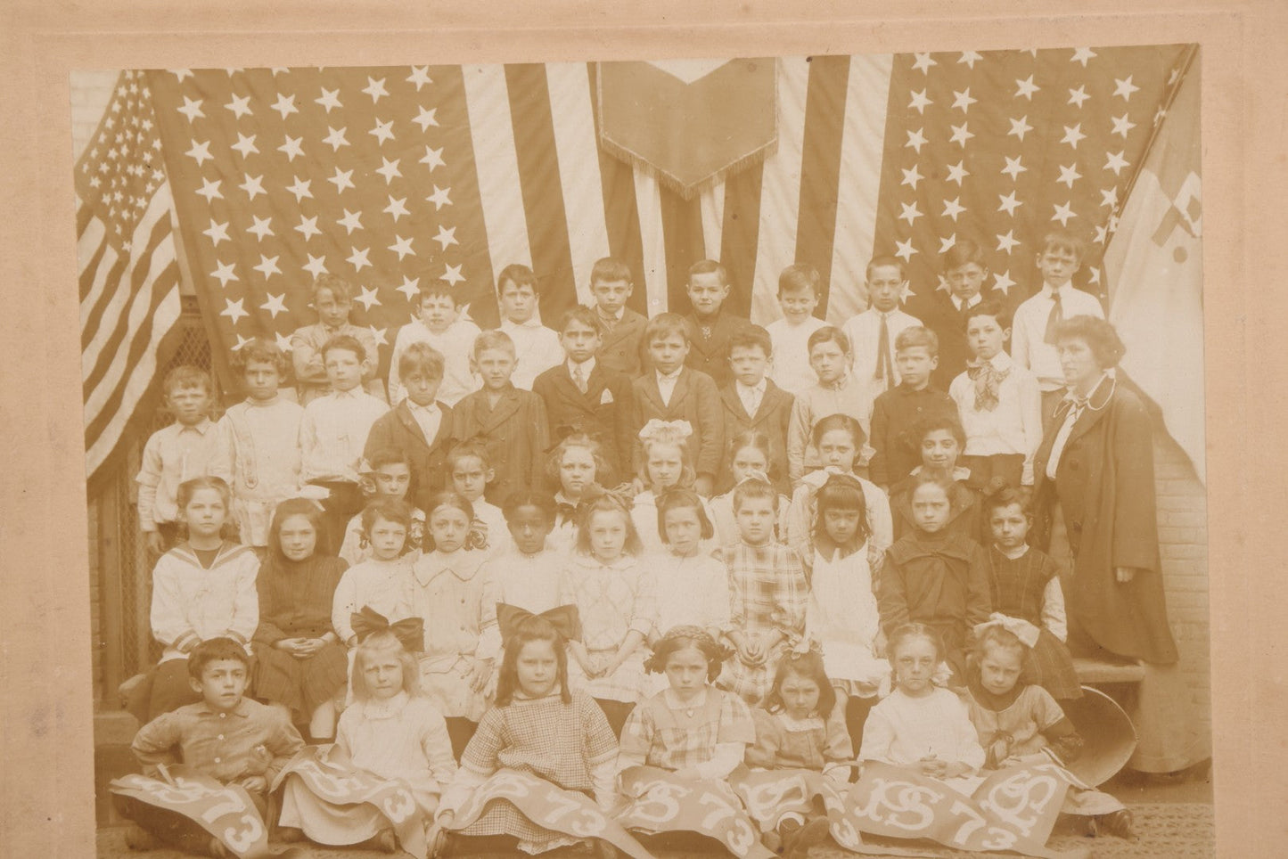 Lot 069 - Antique Boarded Photograph Of An Integrated Elementary School Class, Posing With Large 48 Star American Flag Bunting And Patriotic Decor, Pennants From P.S. 73, Teacher In Photo, Circa 1895