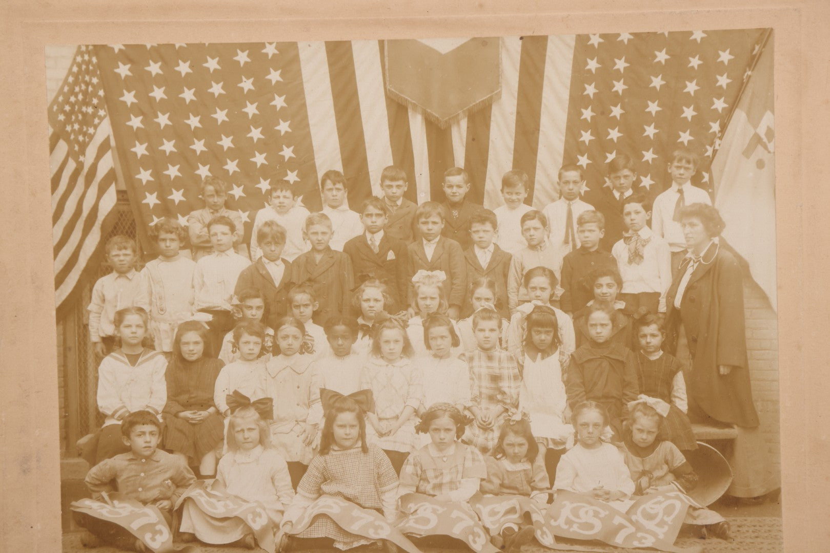 Lot 069 - Antique Boarded Photograph Of An Integrated Elementary School Class, Posing With Large 48 Star American Flag Bunting And Patriotic Decor, Pennants From P.S. 73, Teacher In Photo, Circa 1895