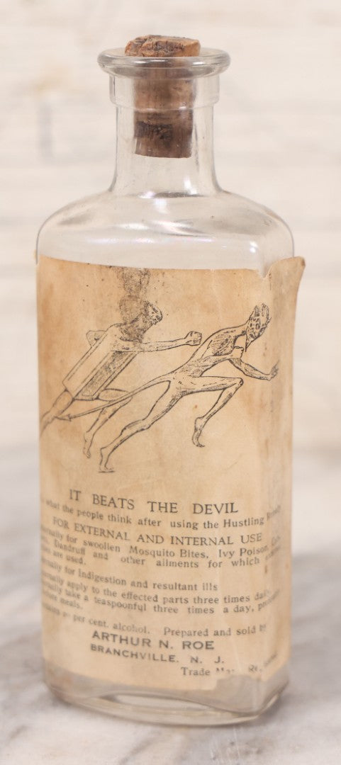 Lot 060 - Antique Clear Glass Bottle With Paper Label “It Beats The Devil” Showing Devil Race Graphics, Arthur N. Roe, Branchville, New Jersey, 5-1/2" H