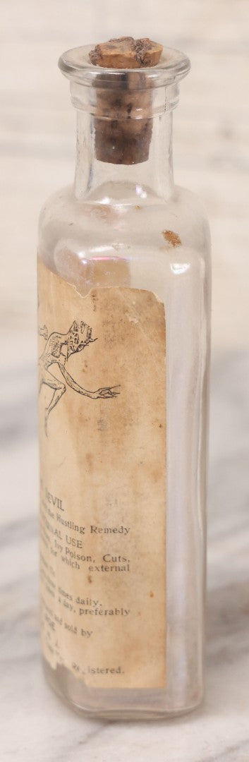 Lot 060 - Antique Clear Glass Bottle With Paper Label “It Beats The Devil” Showing Devil Race Graphics, Arthur N. Roe, Branchville, New Jersey, 5-1/2" H