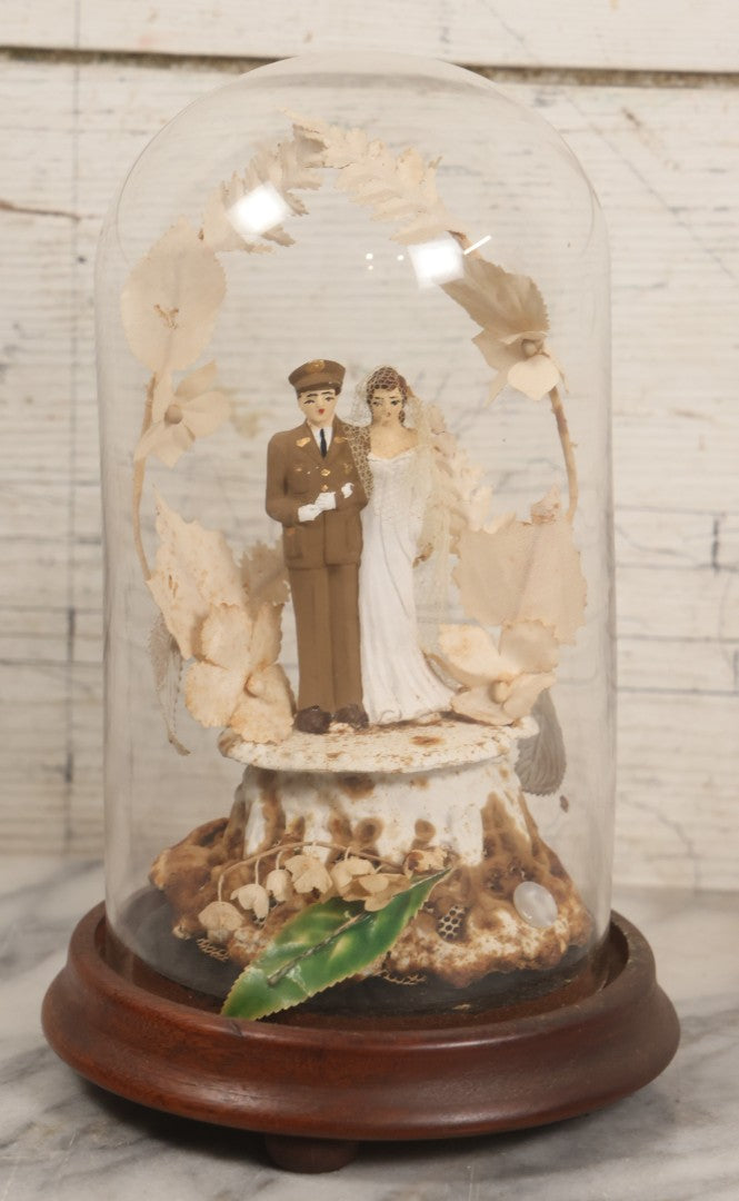 Lot 035 - Vintage Wwii Era Servicemember Wedding Cake Topper In Glass Dome Cloche With Bride, Groom In Military Uniform, Faux White Flowers, And Wood Base, 9-1/2" H