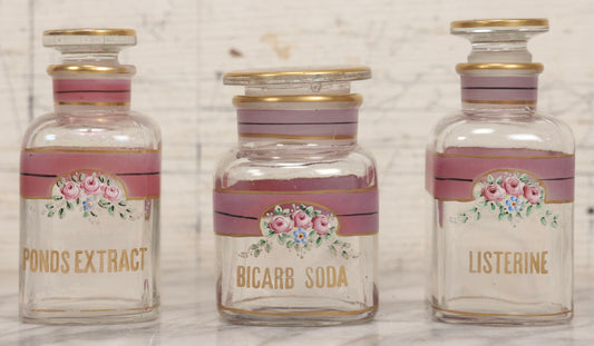 Lot 051 - Antique Three-Piece Painted Glass Bathroom Apothecary Jar Set For Listerine, Bicarb Soda, And Ponds Extract With Gold Lettering, Decorative Lines, And Matching Rose-Decorated Stoppers
