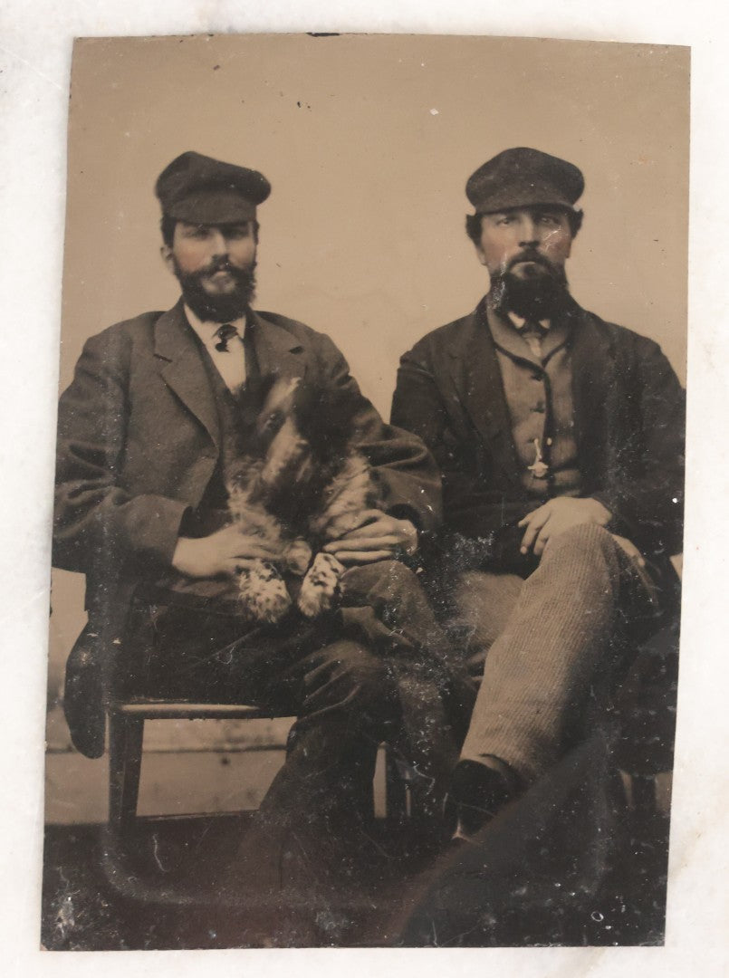 Lot 111 - Antique Tintype Photograph Of Two Bearded Men In Caps With Rambunctious Spotted Dog Seated On Lap, Dog Blurred From Motion