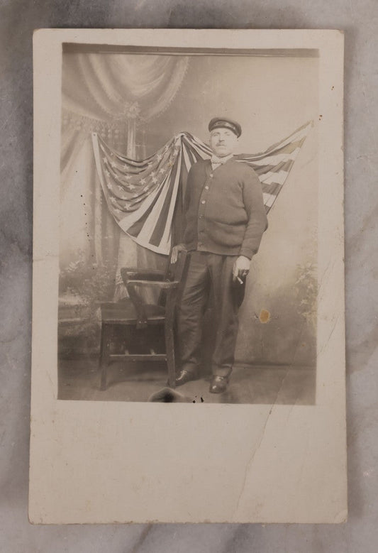 Lot 174 - Antique Real Photo Postcard R.P.P.C. Of Man In Uniform And Cap With Cigarette Before American Flag Backdrop, Circa 1910s