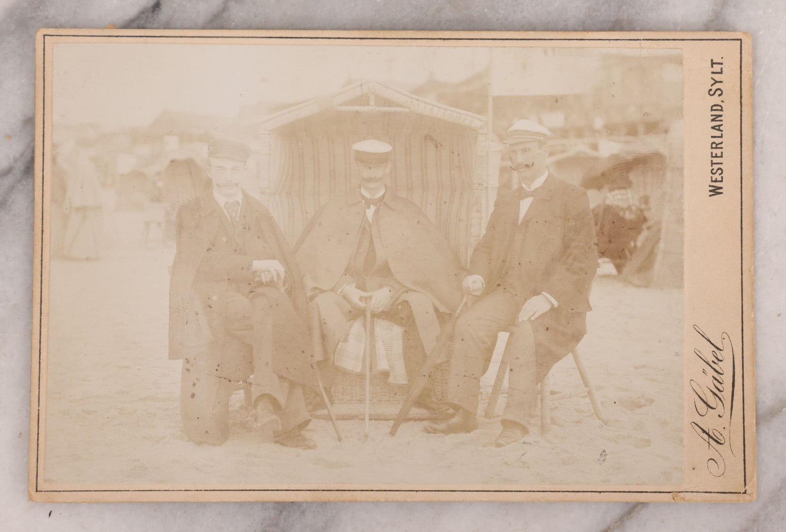 Lot 179 - Antique Board-Mounted Photograph Of Three Mustachioed Men Posed On Beach In Suits With Capes And Walking Sticks Before Striped Beach Windbreak, Westerland, Sylt, Photographed By A. Gabel