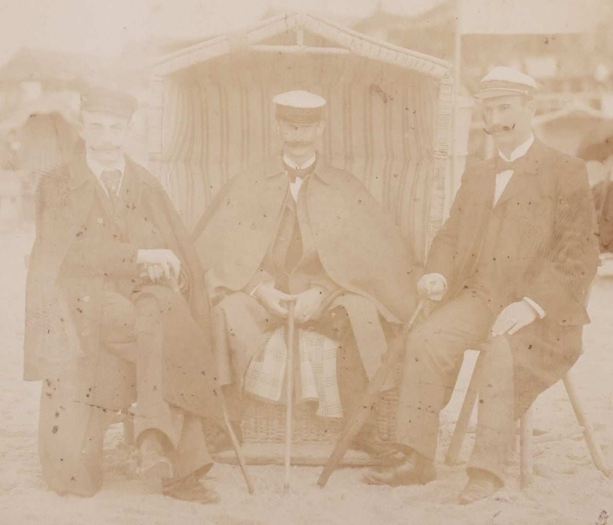 Lot 179 - Antique Board-Mounted Photograph Of Three Mustachioed Men Posed On Beach In Suits With Capes And Walking Sticks Before Striped Beach Windbreak, Westerland, Sylt, Photographed By A. Gabel