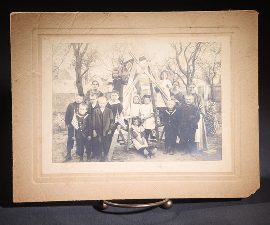 Lot 137 - Antique Board-Mounted Photograph Of A Group Of Children With American Flags Posed Around A Wooden Structure Outdoors